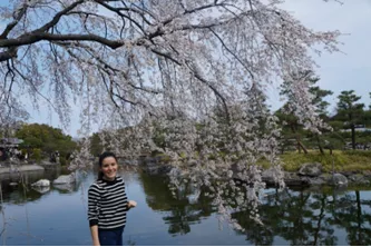 Young woman stands in front of spring lake setting under a cherry blossom tree dressed in striped sweater smiling