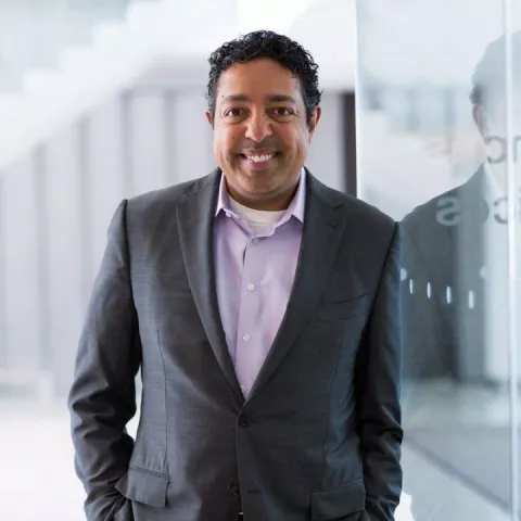 A smiling man in grey blazer and light purple dress shirt smiles while leaning against a glass surface