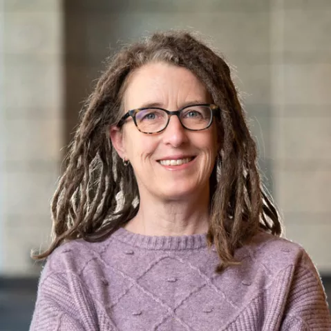 A woman with medium length hair, purple sweater smiles with glasses against a blury grey building background