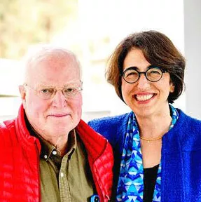 An older man in a red jacket stands smiling next to a smiling woman with short brown hair in glasses and a blue jacket