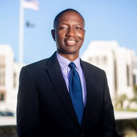 A smiling man in navy blazer with dress shirt and tie stands in front of buildings with flags
