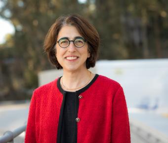 Woman with short brown hair and glasses smiles dressed in red and black sweater against an outdoor setting