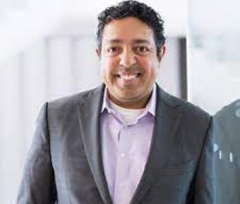 A man in grey suit and lavender business shirt stands against a blurry grey office setting