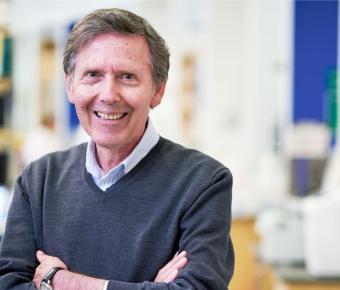 Man with grey hair smiles and stands with hands crossed dressed in blue collared shirt and grey v-neck sweater against a blurry lab setting