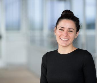 A smiling young woman with dark brown hair in a bun with a black long-sleeve shirt stands in front of blurry grey office space