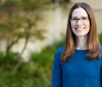 A professional headshot of a smiling woman with long, light brown hair in a blue top against an outdoor blurry setting
