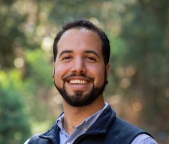 Smiling young man with beard and dark brown hair against a blurry outdoor setting
