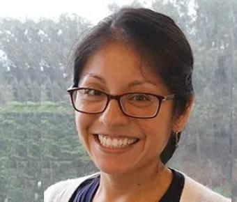 A smiling young woman in glasses with short brown hair poses in front of a blurry window