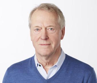 Man with white hair and blue eyes dressed in white collared shirt and v-neck light blue sweater against a white background