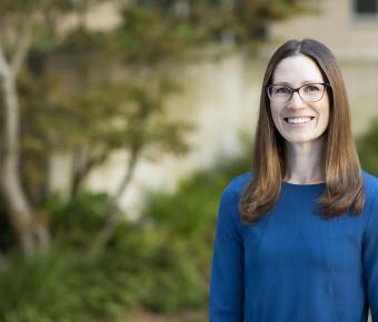 Smiling woman with medium length dark blonde hair and glasses dressed in light blue stands to the right of a blurry outdoor scene