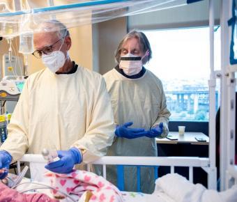 Two healthcare professionals wearing protective gowns, gloves, masks, and face shields attend to a young patient in a hospital bed, surrounded by medical equipment. A window in the background reveals a cityscape.