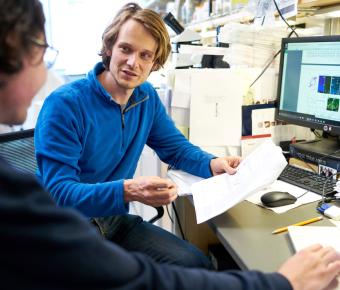 two men sit at a computer in a lab setting holding a paper and in discussion
