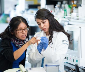 scientist reaches over to fellow female scientist holding clear sample while pointing with finger
