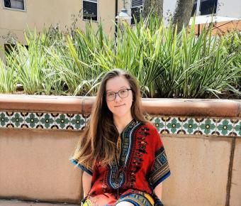 A young woman with long blonde hair sits in colorful dress on terracotta style bench in an outdoor setting