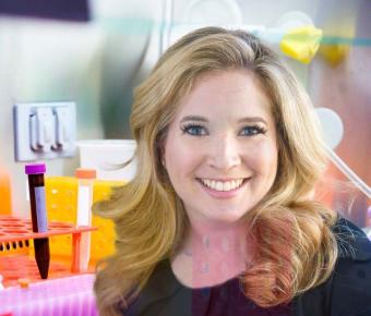 A smiling woman with medium length wavy blonde hair stands in black top against a lab like setting