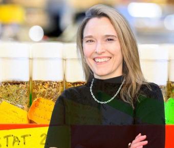 A woman with long blonde hair stands with arms crossed in black sweater in blurry lab setting