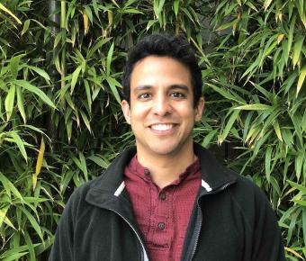 A young man with dark brown hair, red collared shirt and black jacket stands in front of bamboo looking foliage