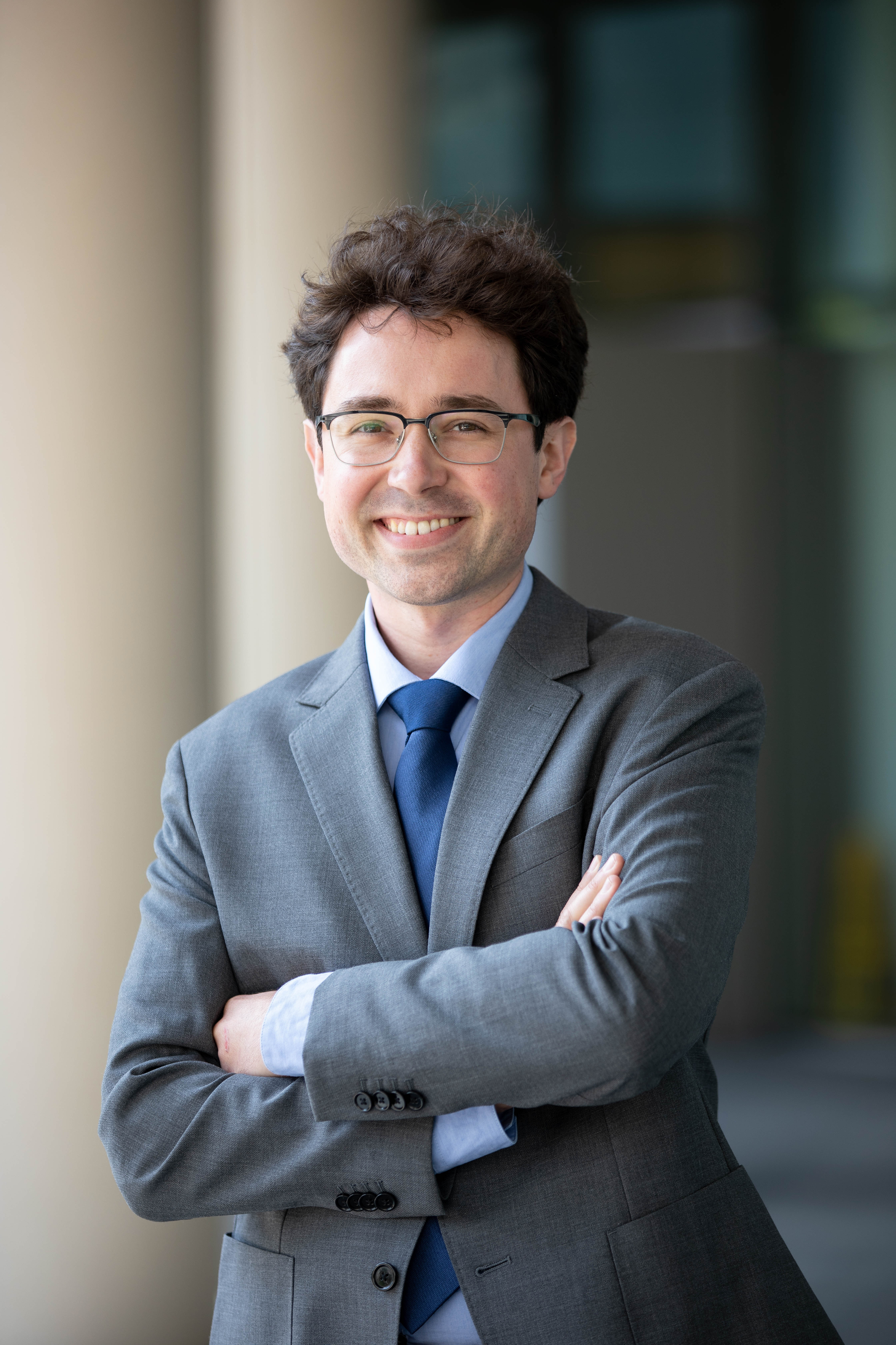 Man in dark suit and tie stands with arms crossed over chest in front of blurry building