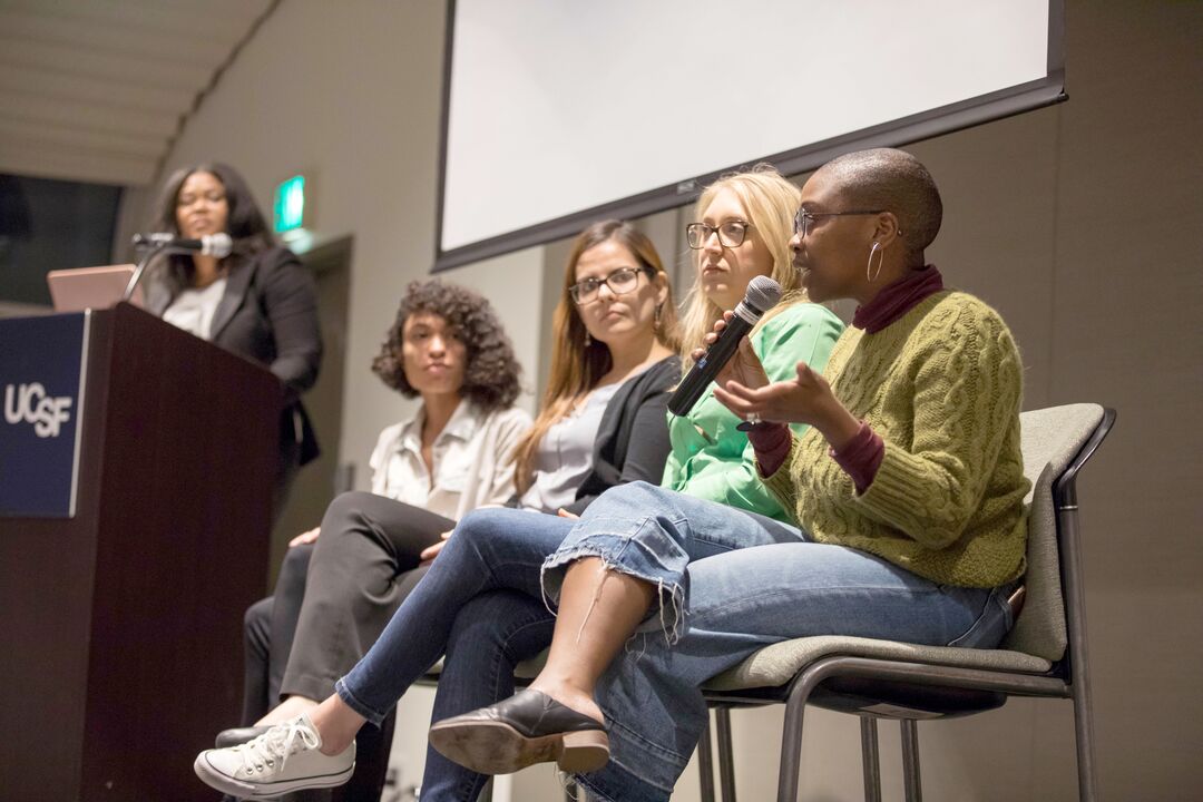 DEI event led by LaMisha Hill who stands at podium on left flanked by four panelists one with microphone responding to inquiryas taken by Susan Merrill